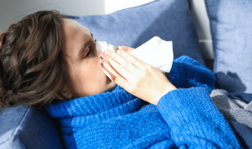 Brown haired woman with blue pullover lies on couch and blows her nose