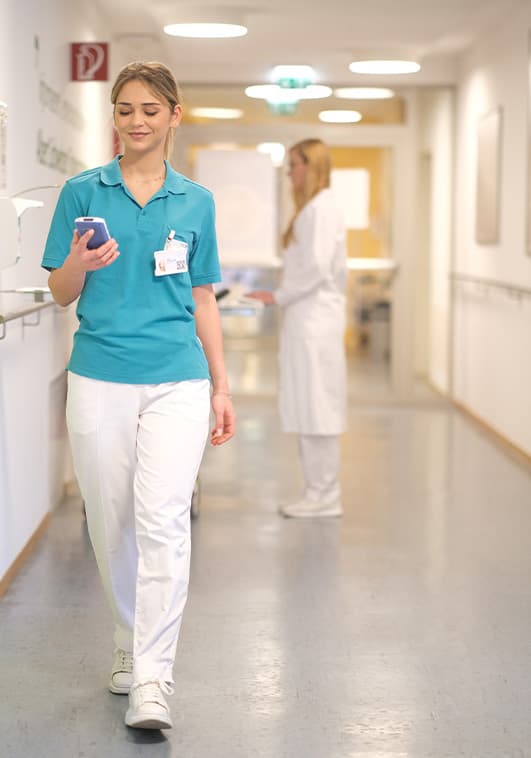Ärztin oder Krankenschwester geht Krankenhauskorridor entlang und blickt dabei auf ihr Handheld-Device von ilviCLINIC, auf dem STEADYTEMP als Drittanbieter-App installiert ist.