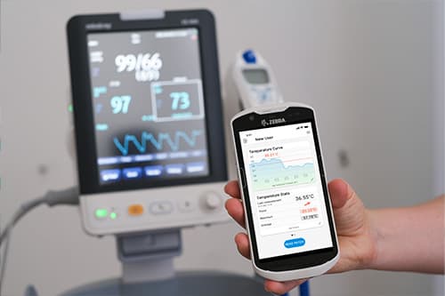 Im Vordergrund das Gerät von ilviCLINIC mit der STEADYTEMP-App darauf – der Temperaturscreen ist geöffnet und man sieht den Temperaturverlauf sowie die Fieberdaten. Im Hintergrund sieht man schemenhaft den Monitor eines medizinischen Geräts.